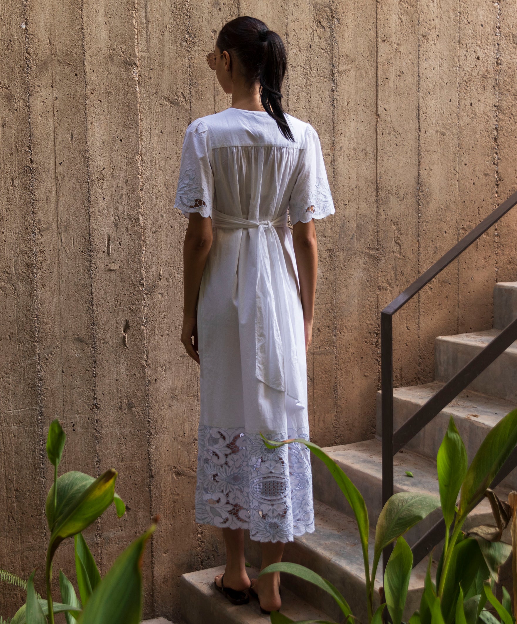 Woman in a white embroidered maxi dress standing on steps with a textured wall and plants in the foreground