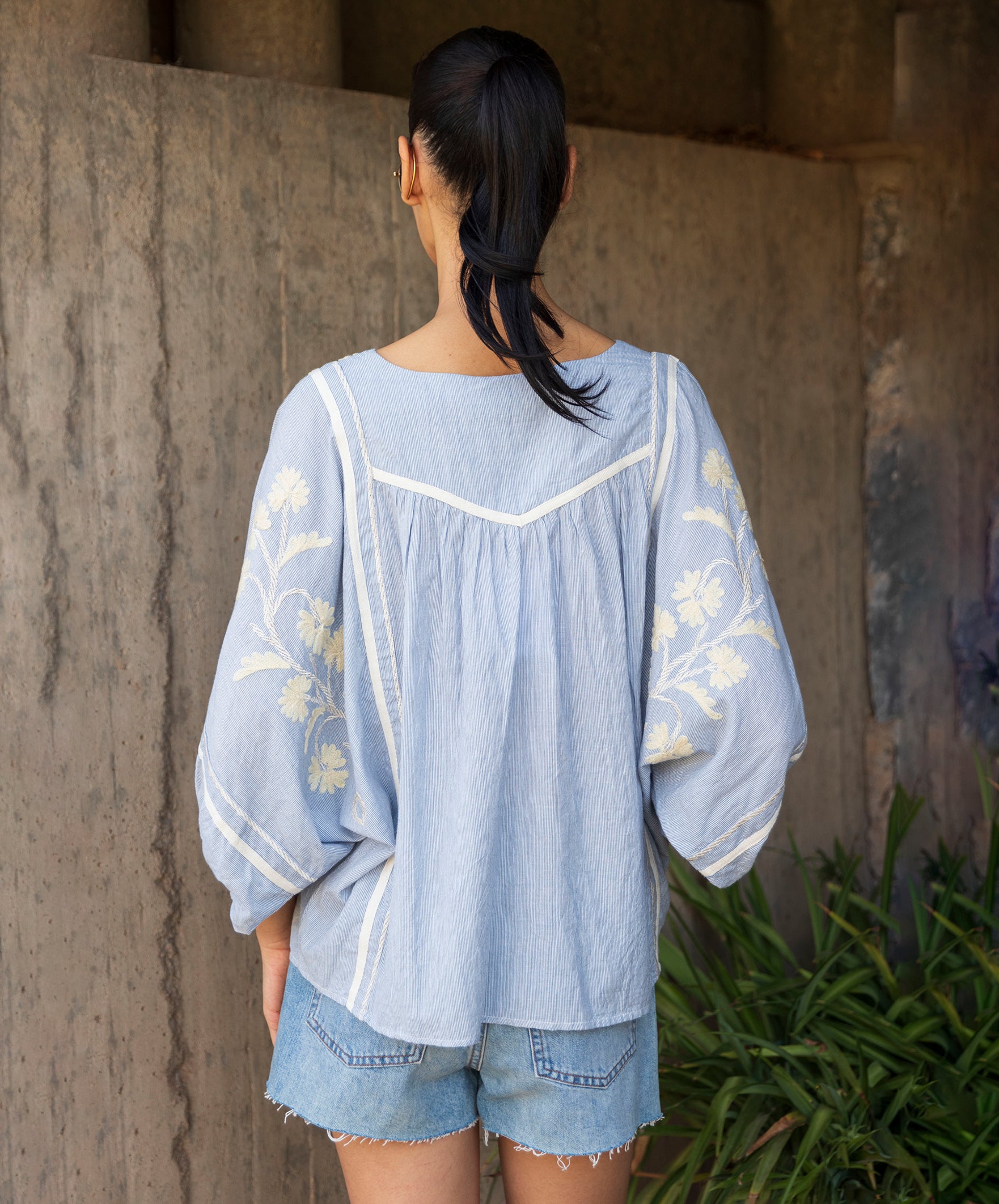 Woman wearing a blue striped embroidered blouse and denim shorts standing against a stone wall.