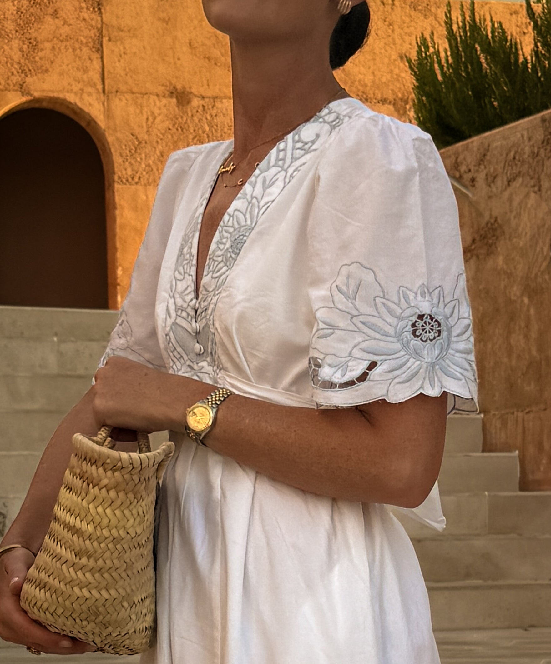 A woman wearing a white embroidered dress with blue floral embroidery on the bodice and sleeve, holding a woven straw bag in front of a warm beige stone wall and steps.