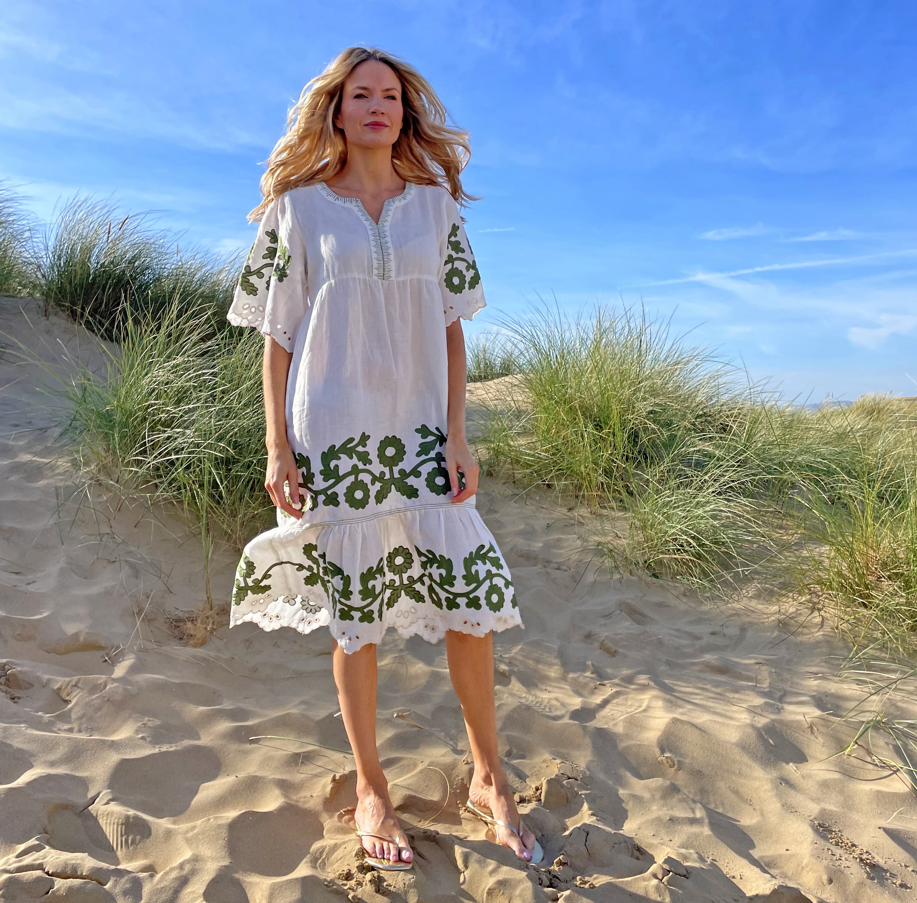 A blond haired model on a beach wearing a white and olive Rose and Rose Agrigento Dress
