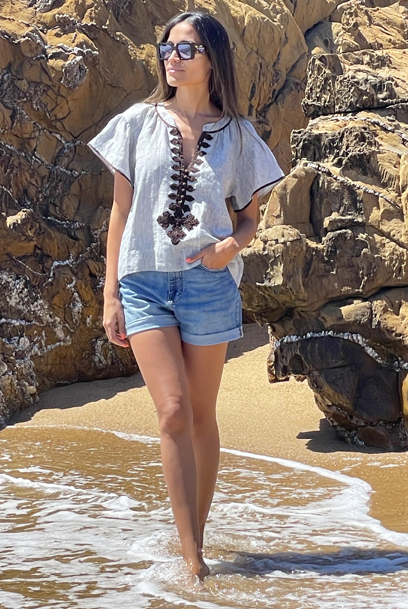 A model walking on a beach wearing a Rose and Rose striped linen Imperia top, denim shorts and sunglasses.