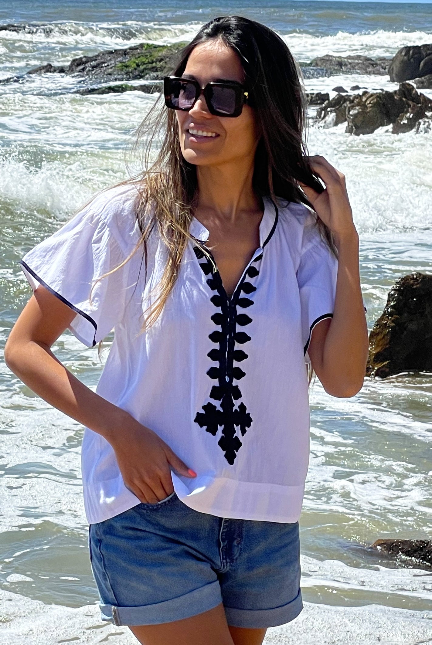  A model stood in front of the sea wearing a Rose and Rose white Imperia top, denim shorts and sunglasses.