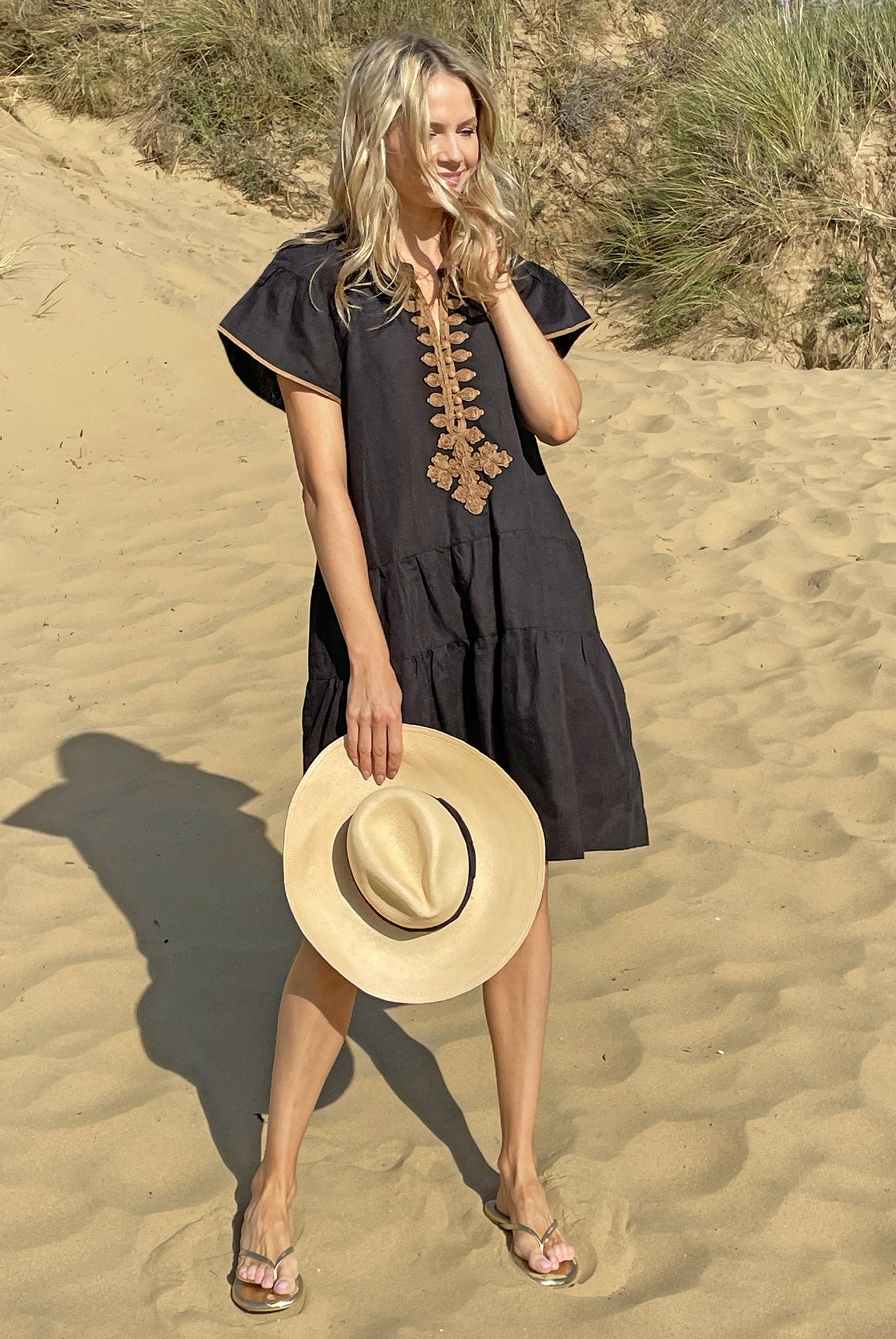 A model on a beach wearing the Rose and Rose Ischia dress in black cotton and holding an Anthony Peto Panama hat.