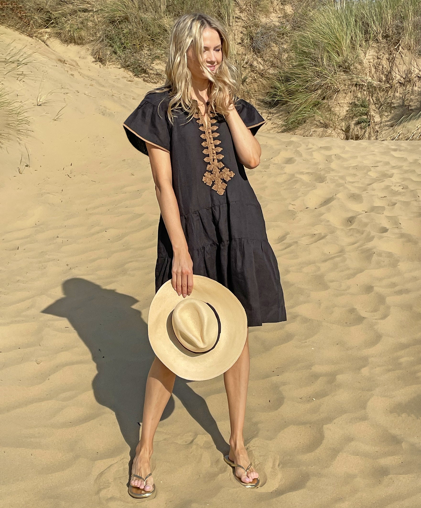 A model on a beach wearing the Rose and Rose Ischia dress in black cotton and holding an Anthony Peto Panama hat.
