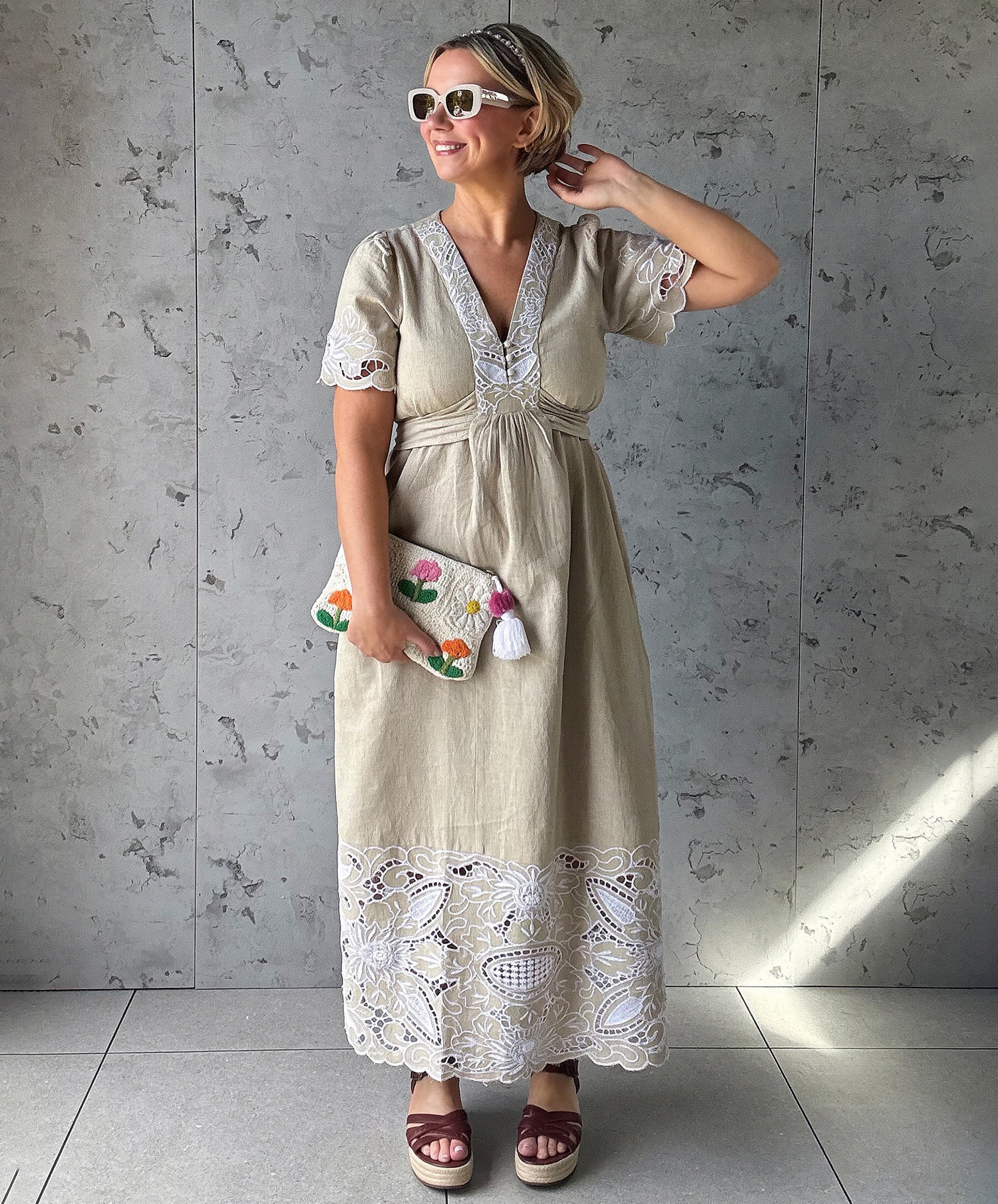 Influencer @rosemaejewel wearing a Hawthorn natural dress with lace embroidery details holding a colorful handbag against a gray concrete wall.