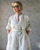 Woman wearing a white dress with green embroidery against a gray wall.