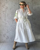 Woman wearing a white dress with green embroidery against a gray concrete wall.