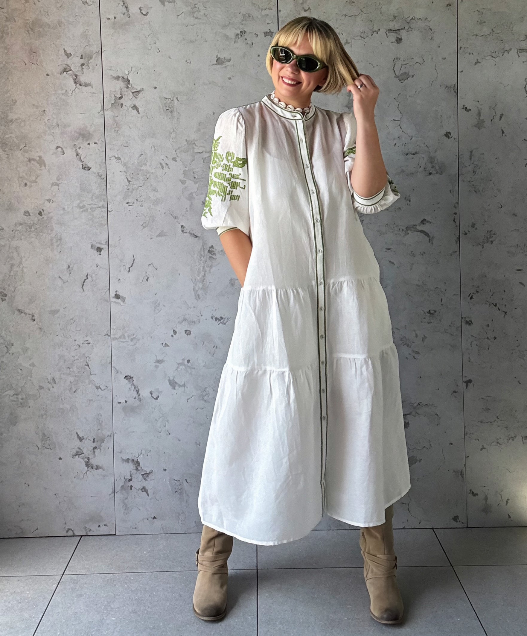 Woman wearing a white dress with green embroidery against a gray concrete wall.