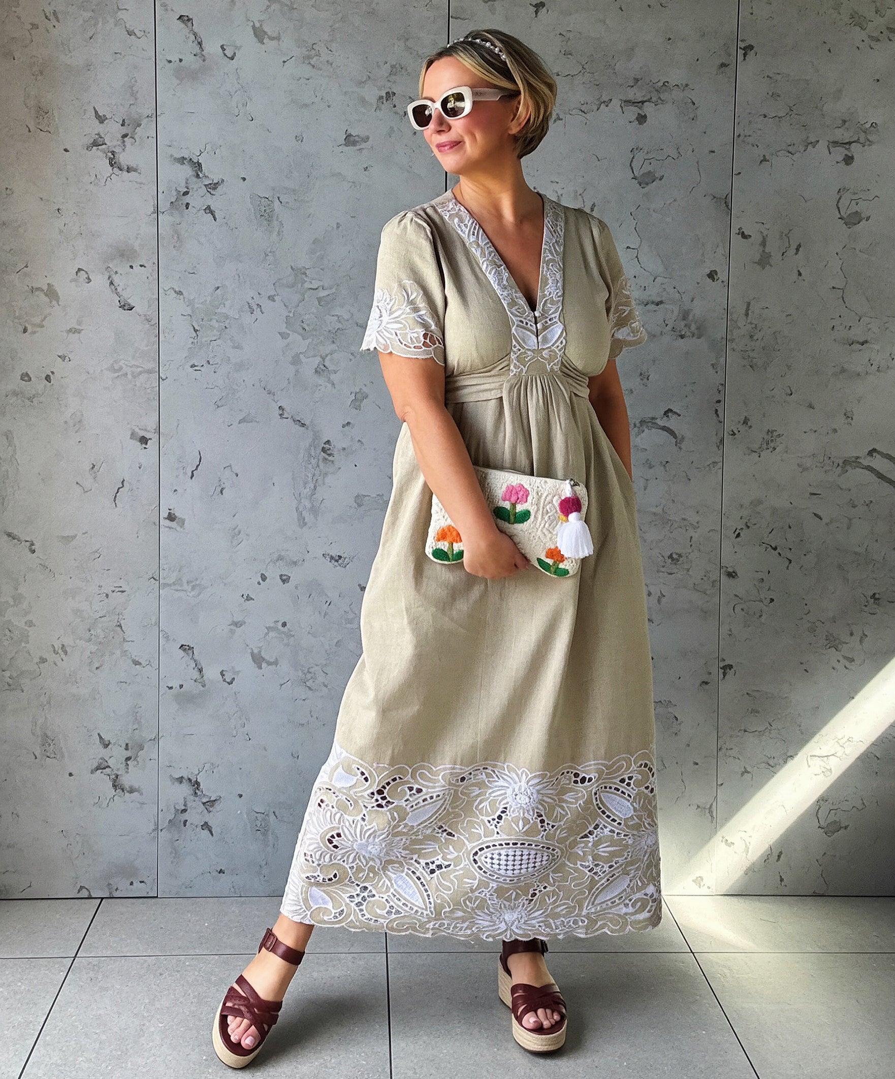 Influencer @rosemaejewel wearing a Hawthorn natural dress with lace embroidery details holding a colorful handbag against a gray concrete wall.