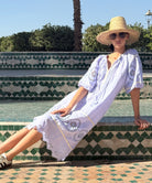 Woman in a blue dress and straw hat sitting on a tiled bench with a pool and palm tree in the background.
