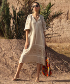 Woman in a natural linen dress with embroidery standing in a desert-like setting with palm trees.