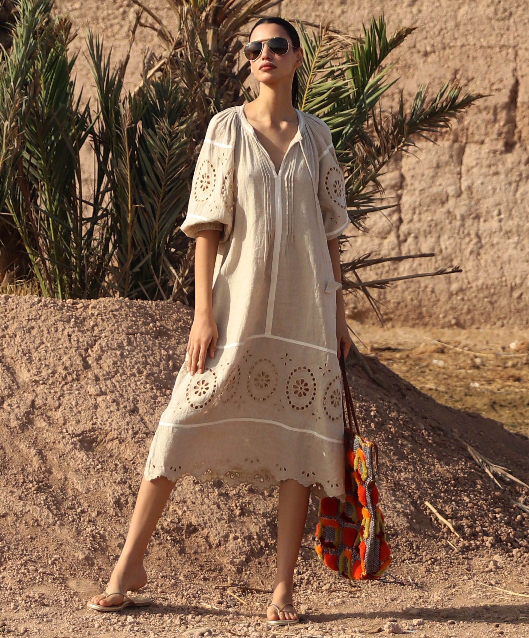 Woman in a natural linen dress with embroidery standing in a desert-like setting with palm trees.