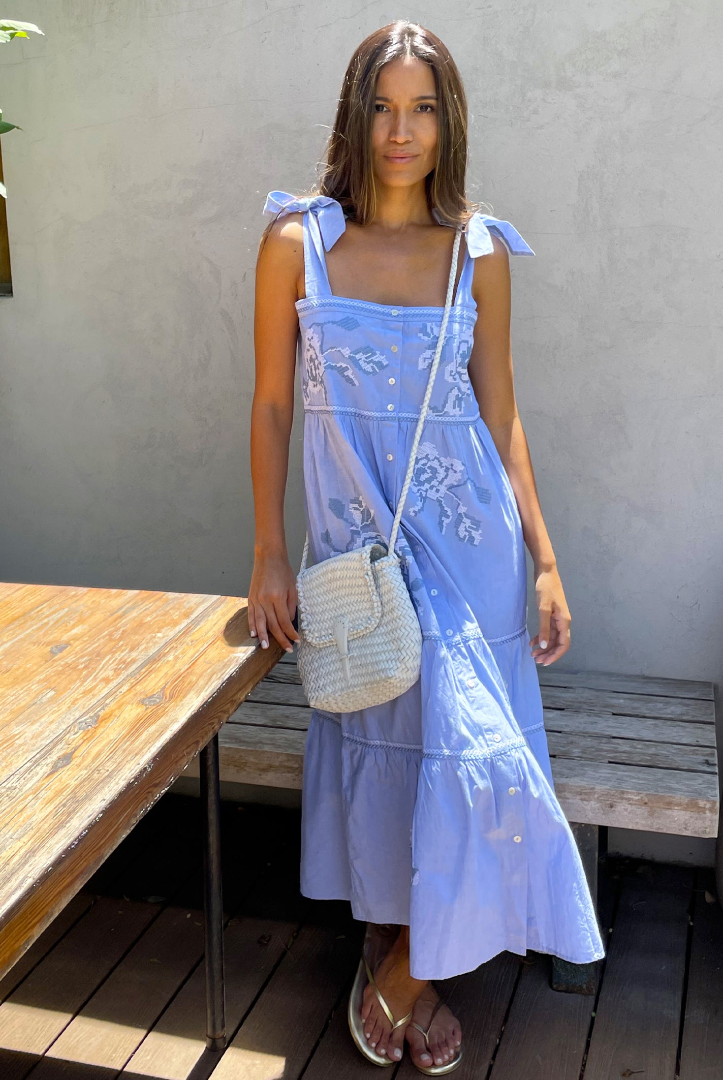 A model stood in a garden wearing a Rose and Rose blue cotton Marina sundress and a white leather Dragon bag.