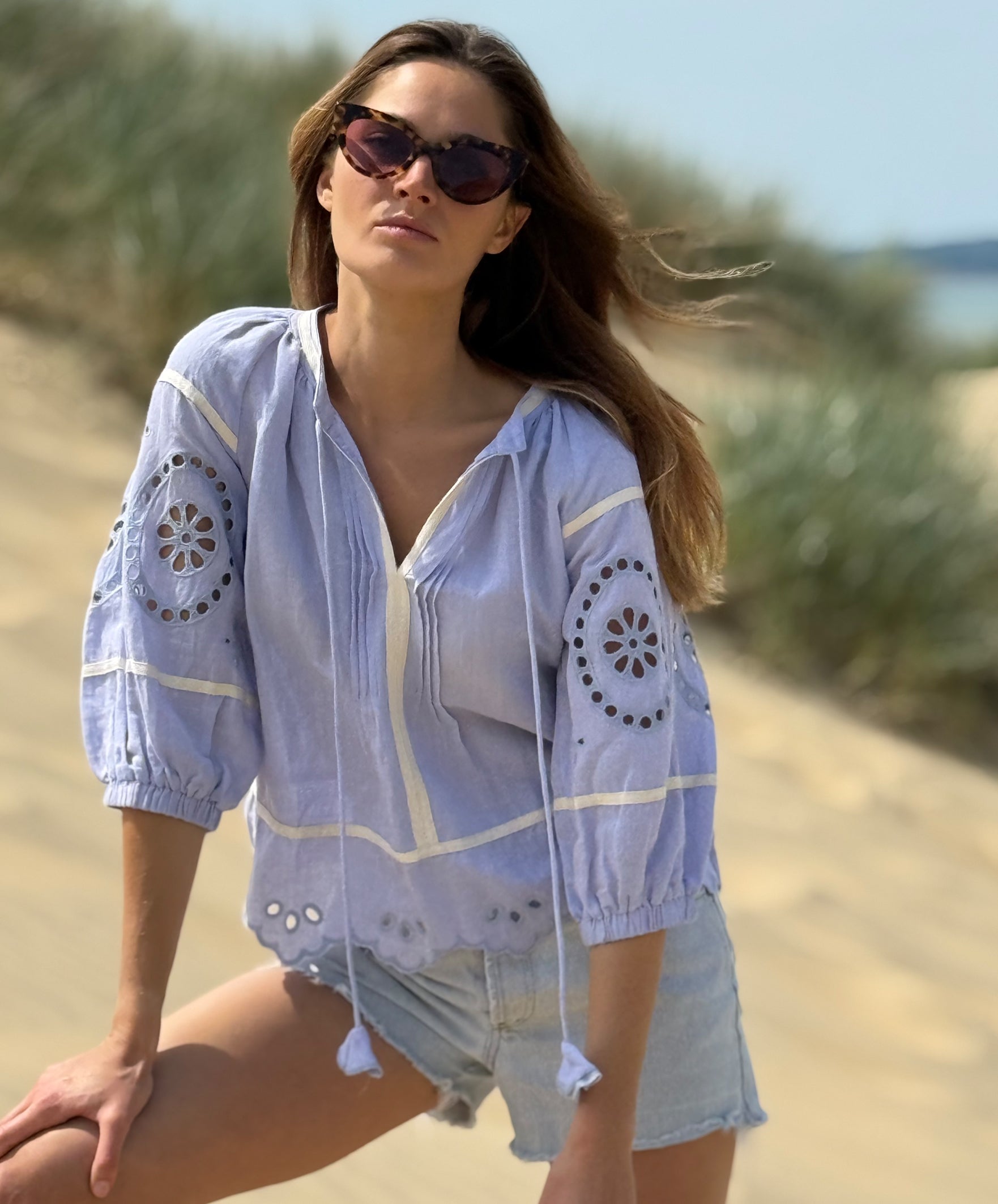 Woman in a light blue floral embroidered linen top on a sandy beach.