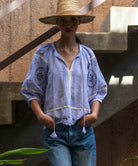 Woman wearing a light blue embroidered top and straw hat standing against a textured wall.