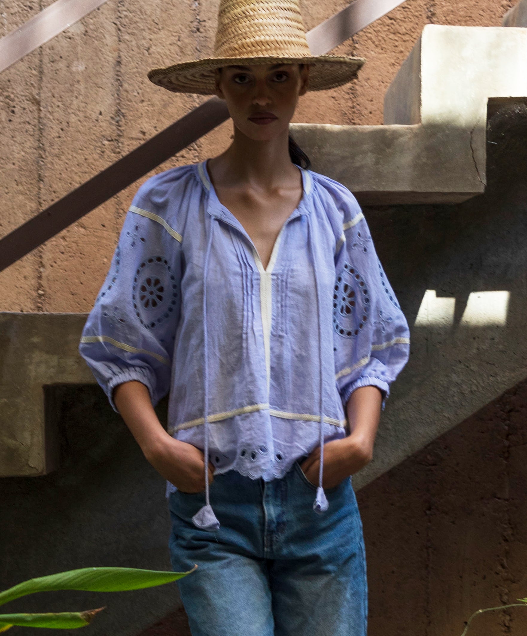 Woman wearing a light blue embroidered top and straw hat standing against a textured wall.