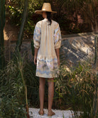 Back view of a woman wearing a floral embroidered gold Oleander dress and straw hat standing in a tropical garden setting