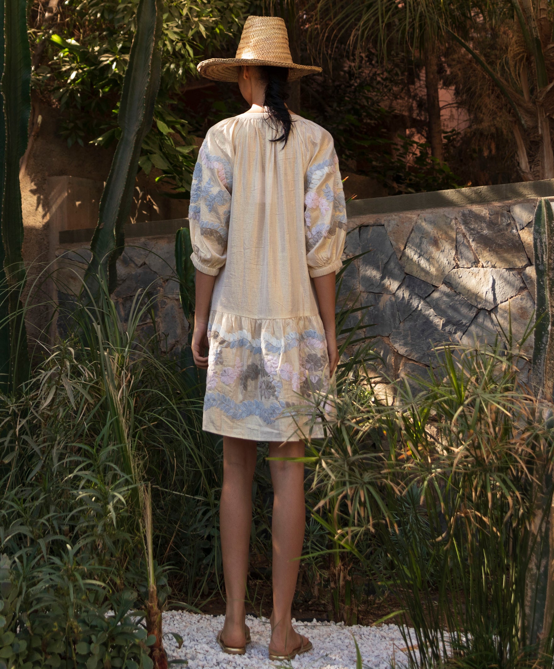 Back view of a woman wearing a floral embroidered gold Oleander dress and straw hat standing in a tropical garden setting