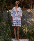 Woman wearing a light pink Oleander dress with blue floral embroidery standing in a tropical garden
