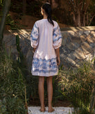 Back view of a woman in a light pink Oleander dress with blue floral embroidery standing in a tropical garden setting.