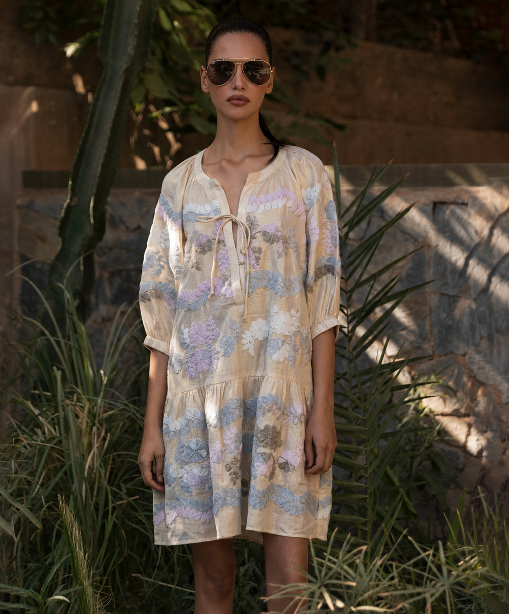 Woman wearing a floral embroidered dress and sunglasses in a natural setting with plants and stone wall.