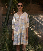 Woman wearing a floral embroidered dress and sunglasses in a natural setting with plants and stone wall.