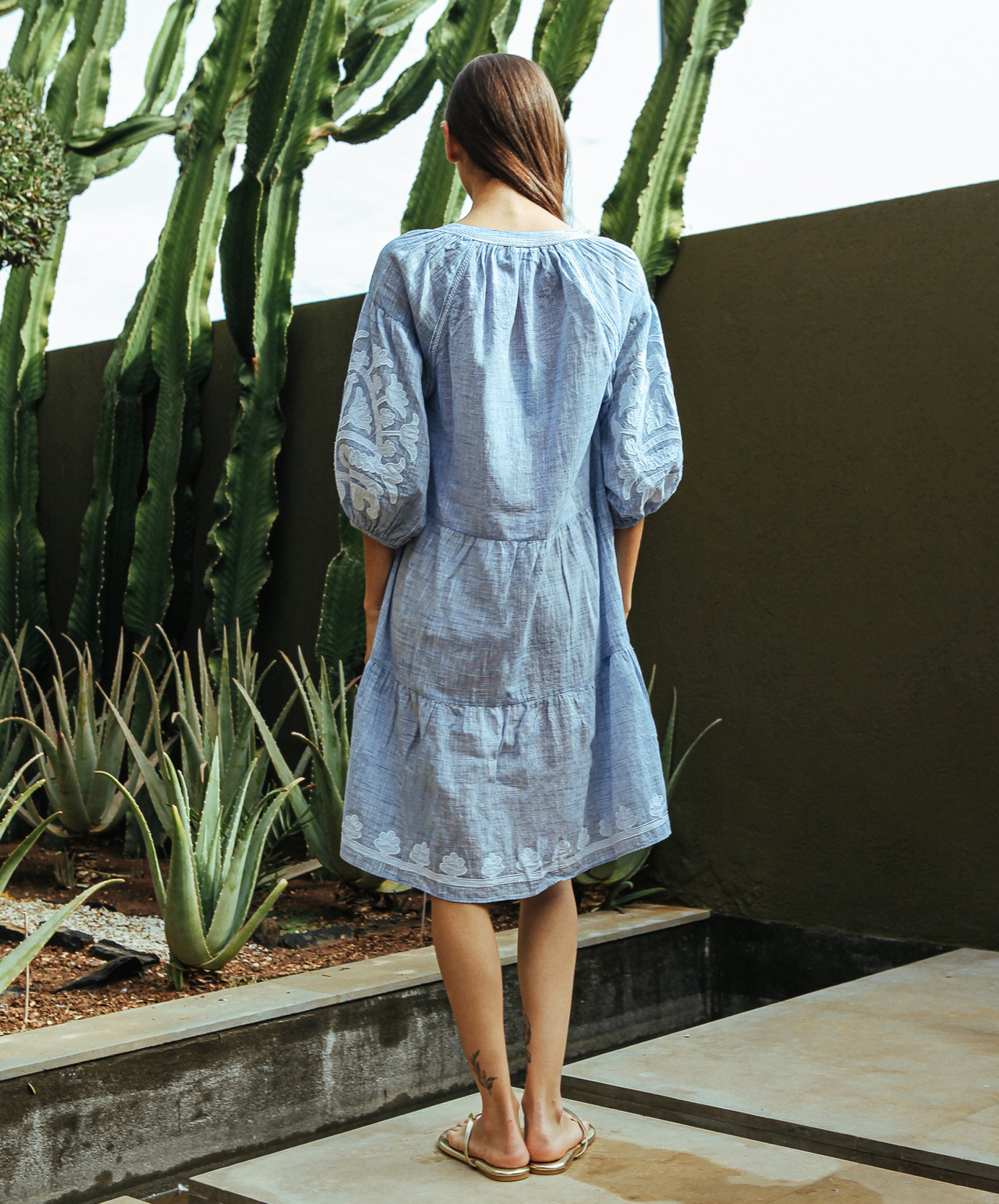 A back view of a model stood in a cacti garden wearing a Rose and Rose Oxford dress in marl blue with white appliqué decoration.