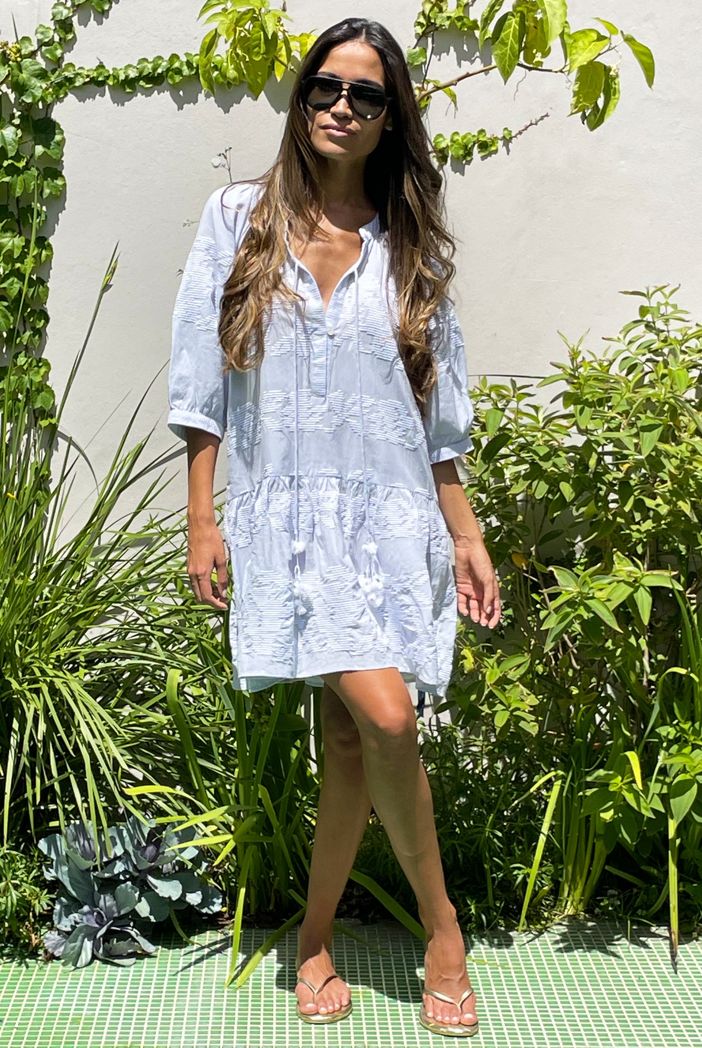 A model stood in a garden wearing sunglasses and a Rose and Rose pale blue cotton embroidered Palermo dress.