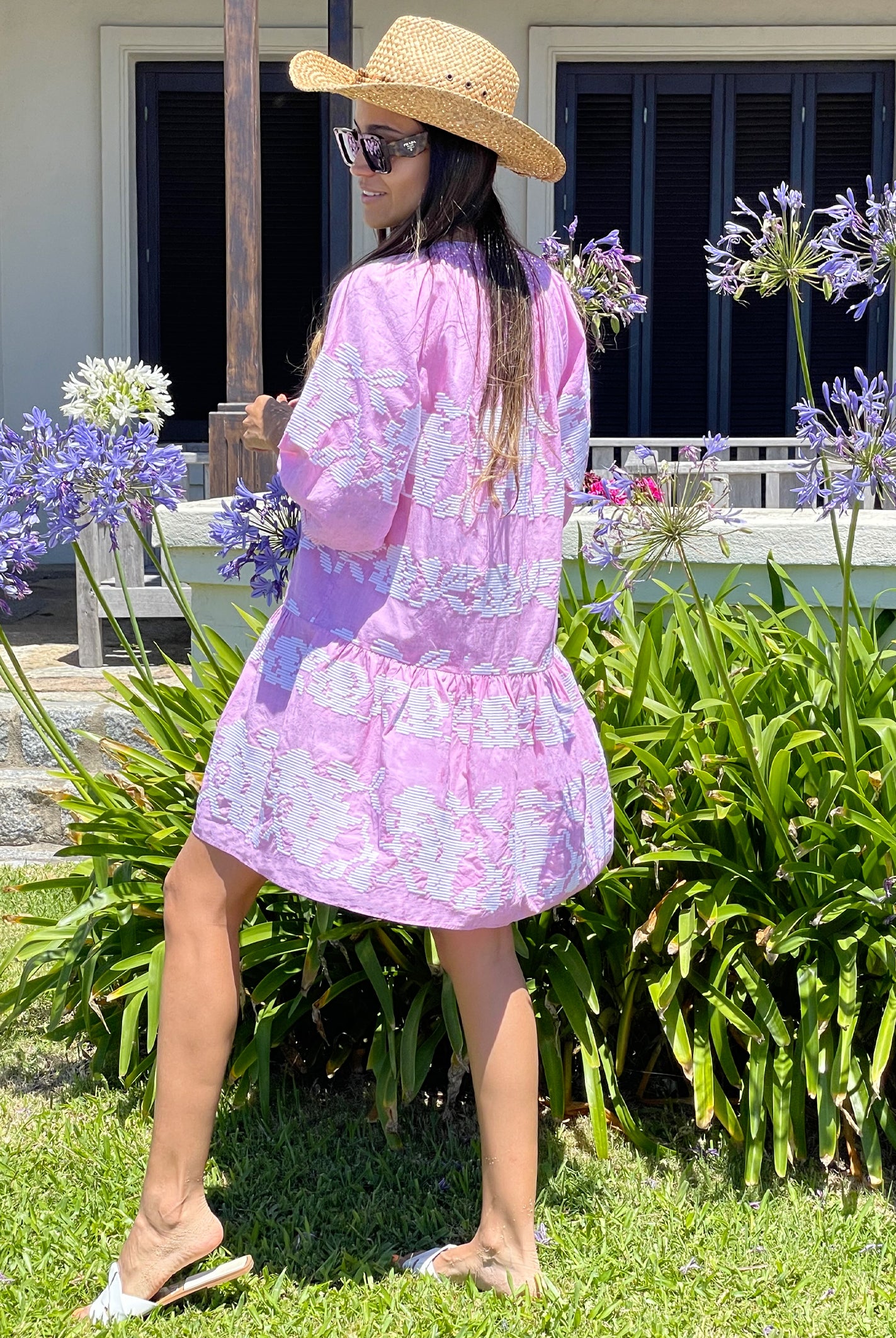 The back view of a model stood in a garden wearing a cowboy hat, sunglasses and a Rose and Rose pale pink Palermo embroidered dress. 