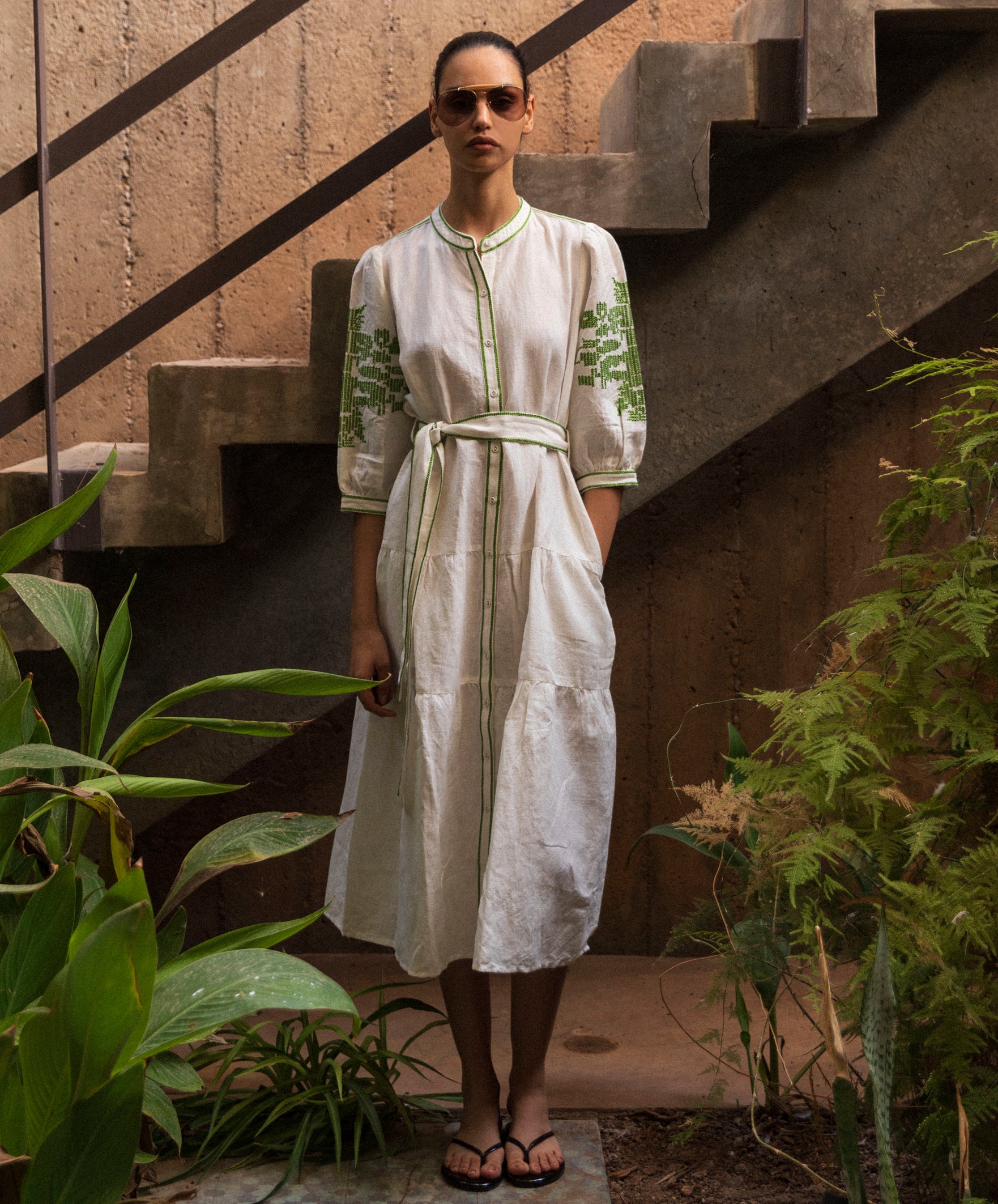 Woman in a white dress with green embroidery standing in a garden setting.