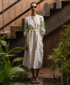 Woman in a white dress with green embroidery standing in a garden setting.