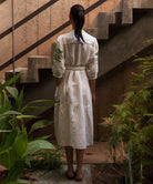 Woman in a white linen dress with olive green embroidery standing on a staircase with plants around
