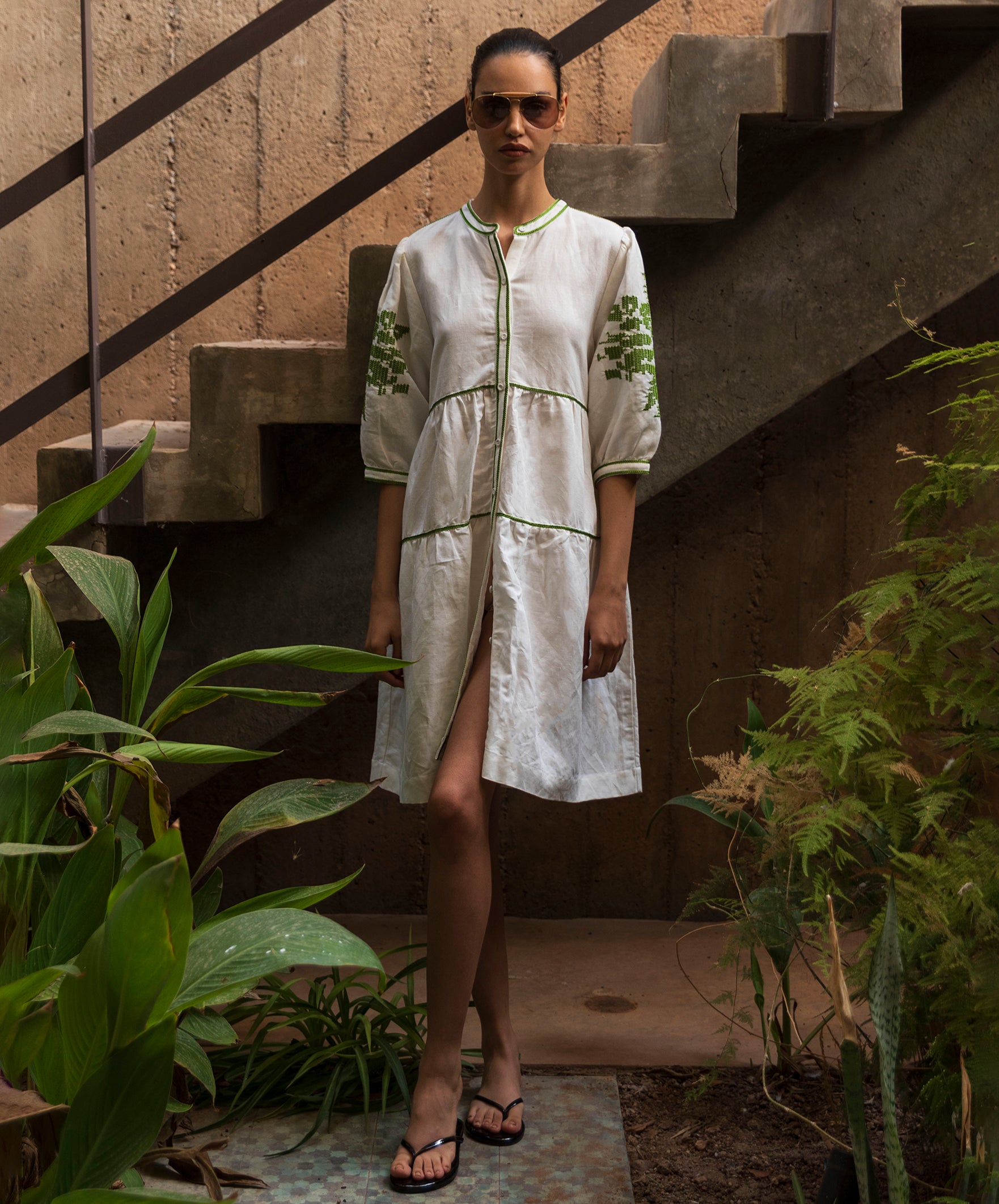 Woman in a white dress with green embroidery standing outdoors near plants and stairs.