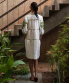 Woman in a white dress with green patterns standing on a staircase surrounded by plants.