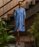 Woman wearing a blue dress with floral patterns standing outdoors near plants and stairs.