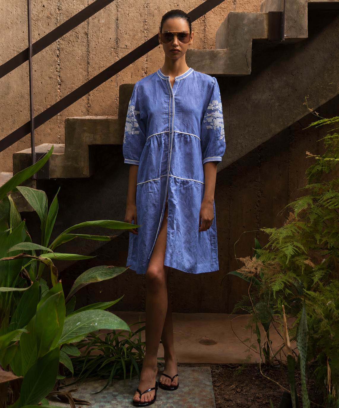 Woman in a blue dress standing outdoors with plants and concrete steps in the background