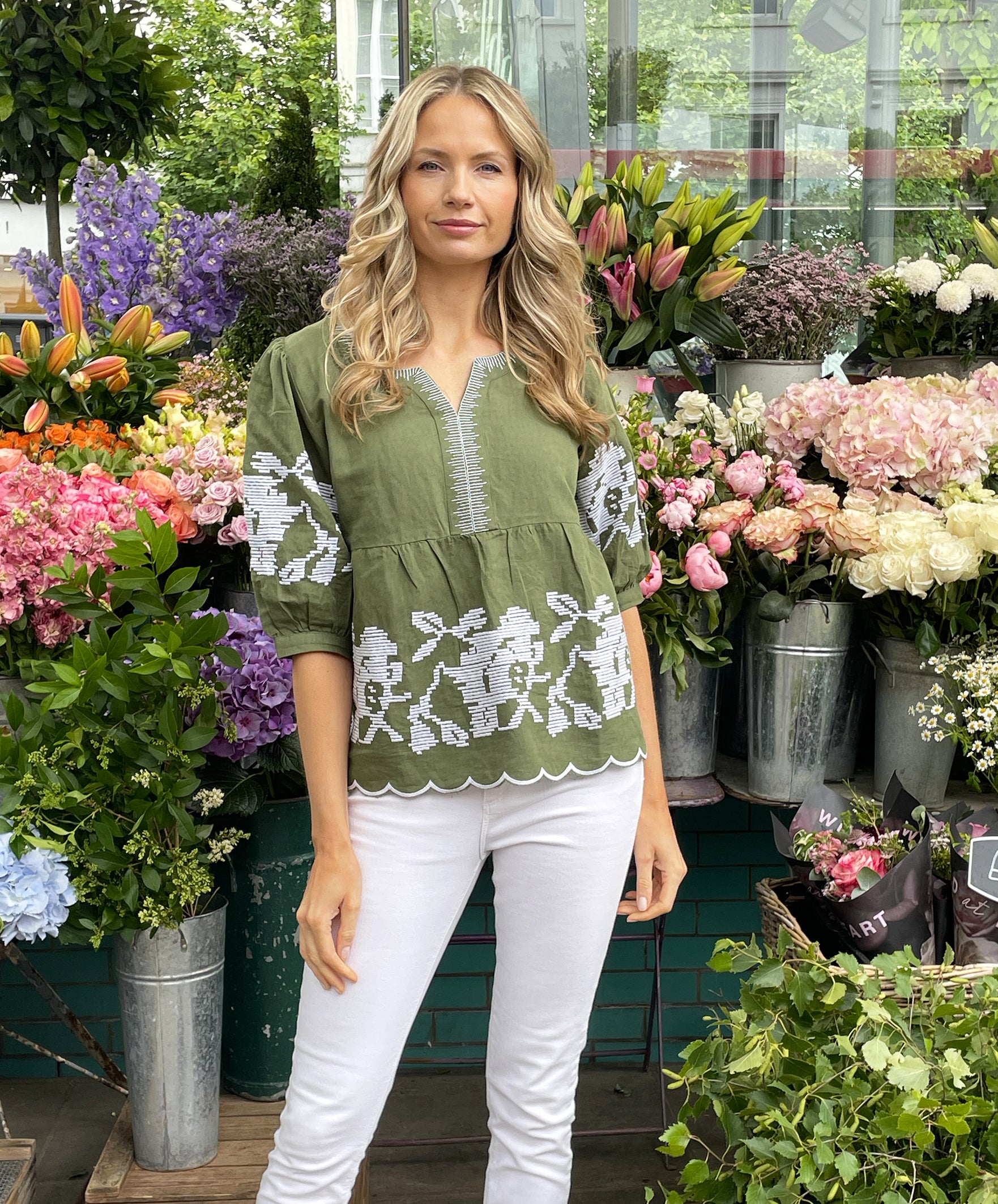 A model surrounded by flowers wearing a Rose and Rose Olive Positano top with white jeans. 