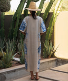 Back view of a woman wearing a long natural Rose dress with blue applique embroidery, standing in a cacti garden.