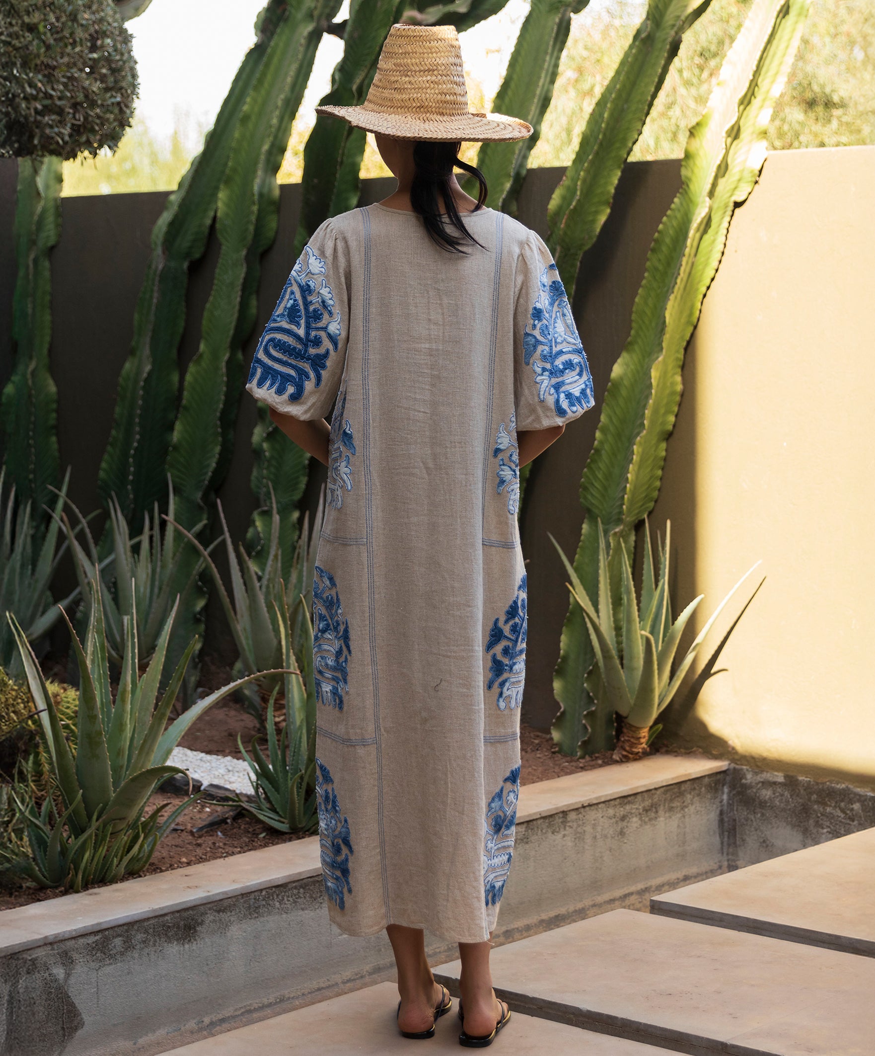 Back view of a woman wearing a long natural Rose dress with blue applique embroidery, standing in a cacti garden.