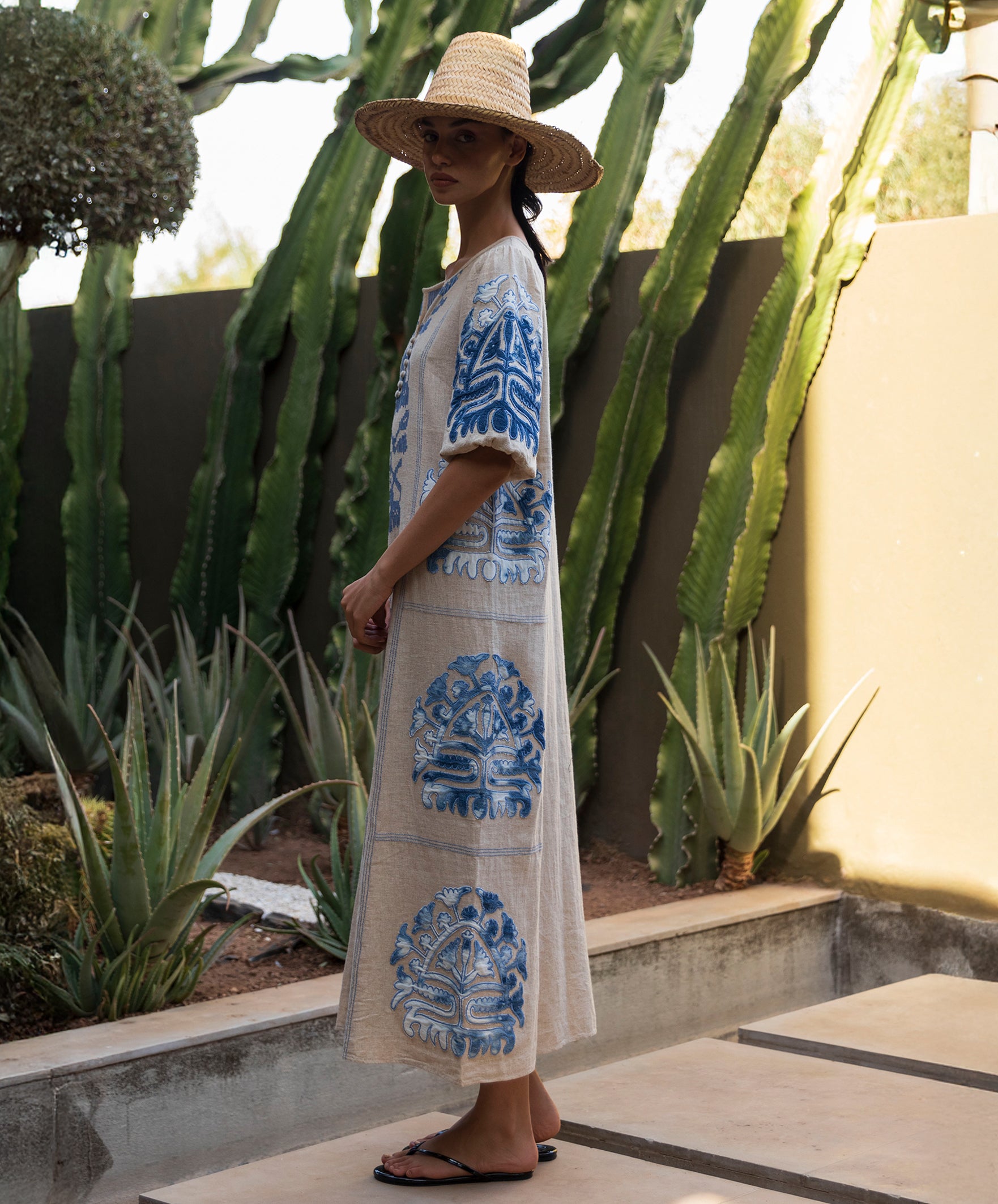 Woman in a long, natural Rose dress with blue applique decoration standing in a garden with cacti.