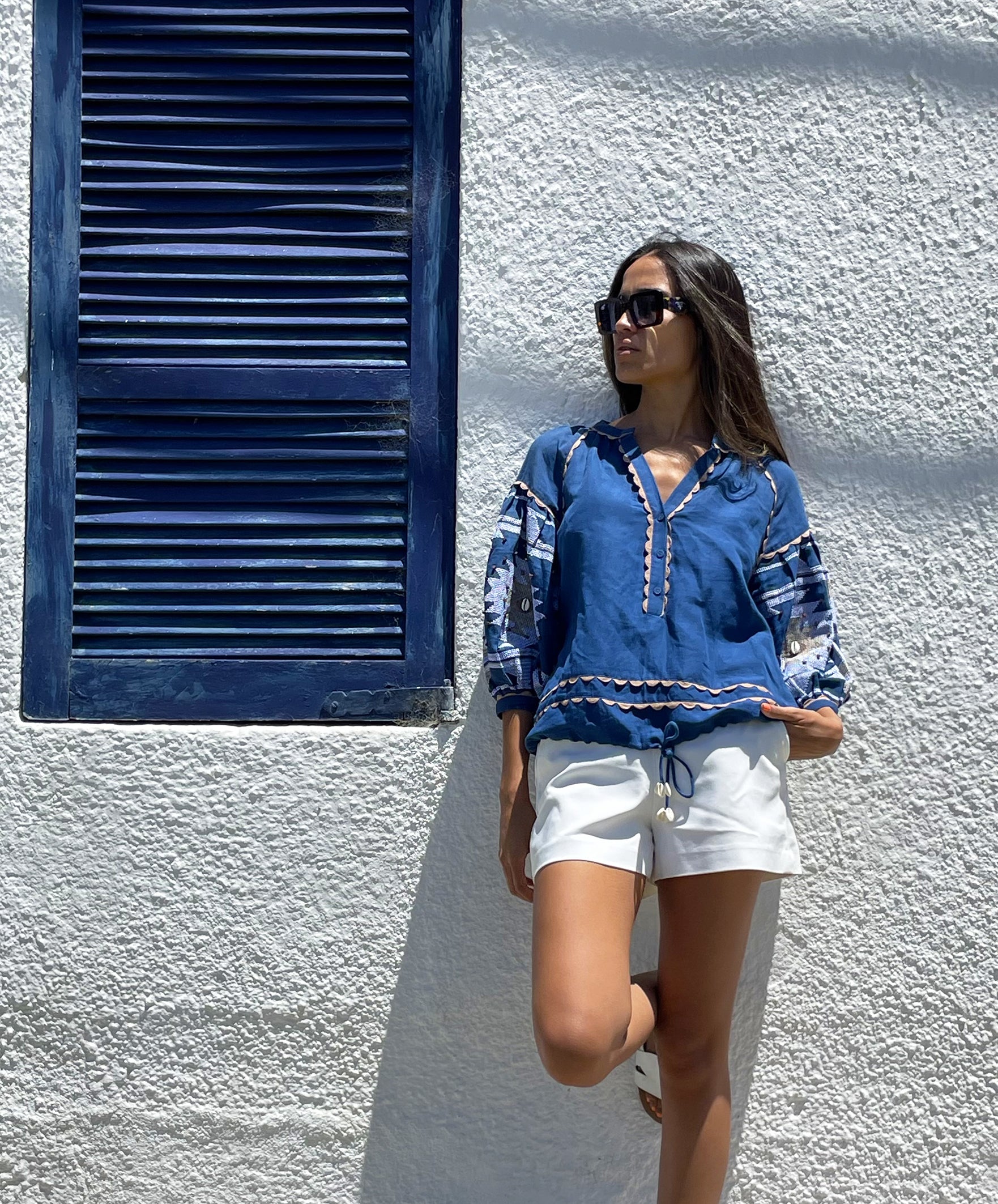 A model stood in front of a white wall wearing a Blue San Marino top, white shorts and sunglasses.