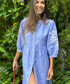 A model in a garden wearing a Rose and Rose blue linen Sicily dress and holding a Dragon Diffusion woven leather bag. 
