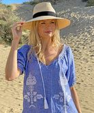 A model on a beach wearing the Rose and Rose Siracusa embroidered blue linen dress and an Anthony Peto Panama hat.