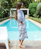 A side view of a model stood in front of a swimming pool wearing a Rose and Rose Snaresbrook striped cotton dress with white embroidery.