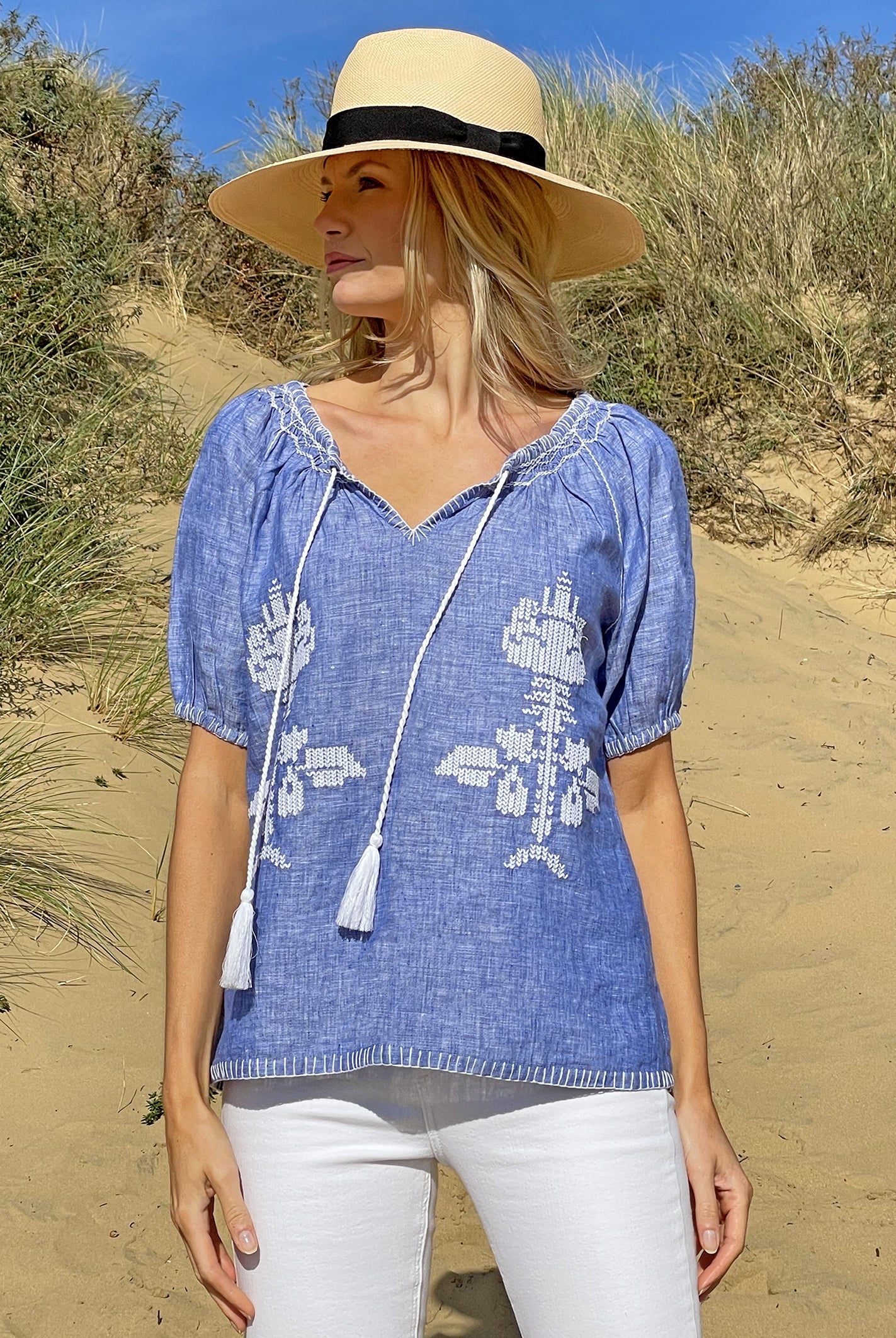 A model on a beach wearing the Rose and Rose Sorrento embroidered blue linen top and an Anthony Peto Panama hat.