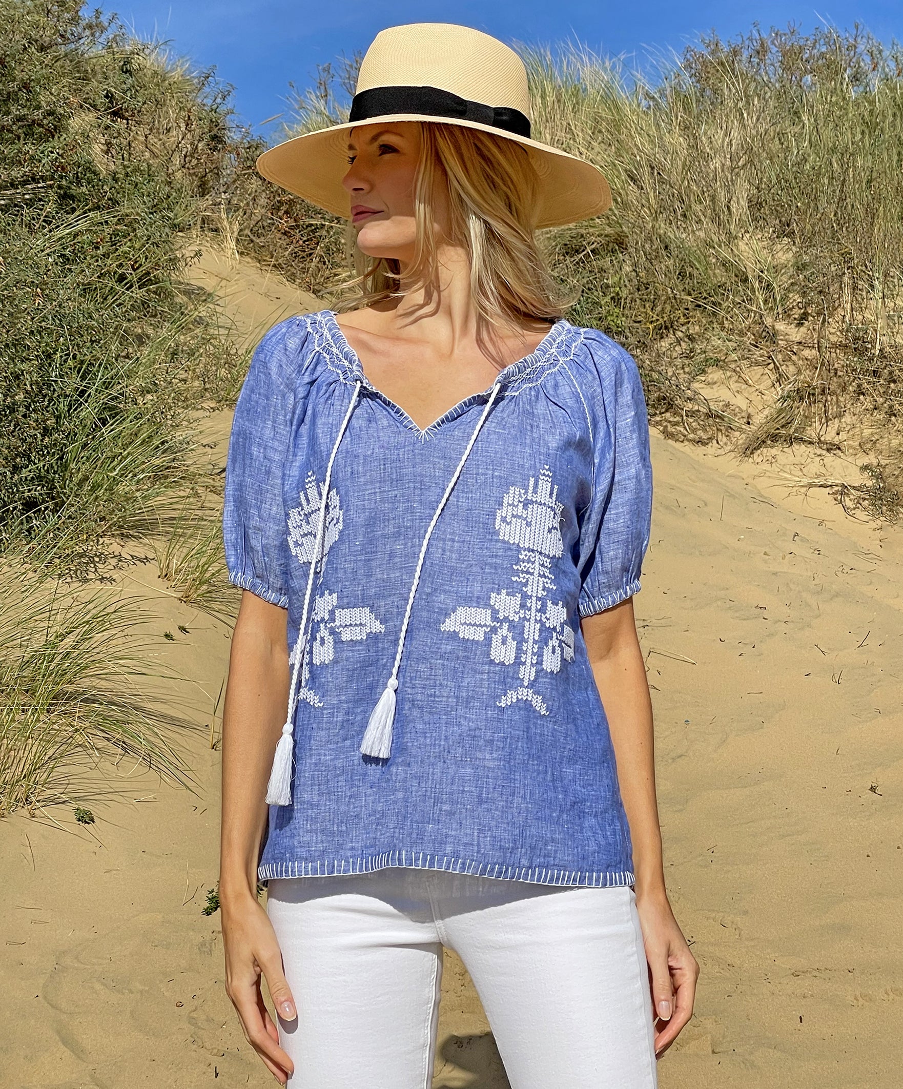 A model on a beach wearing the Rose and Rose Sorrento embroidered blue linen top and an Anthony Peto Panama hat.