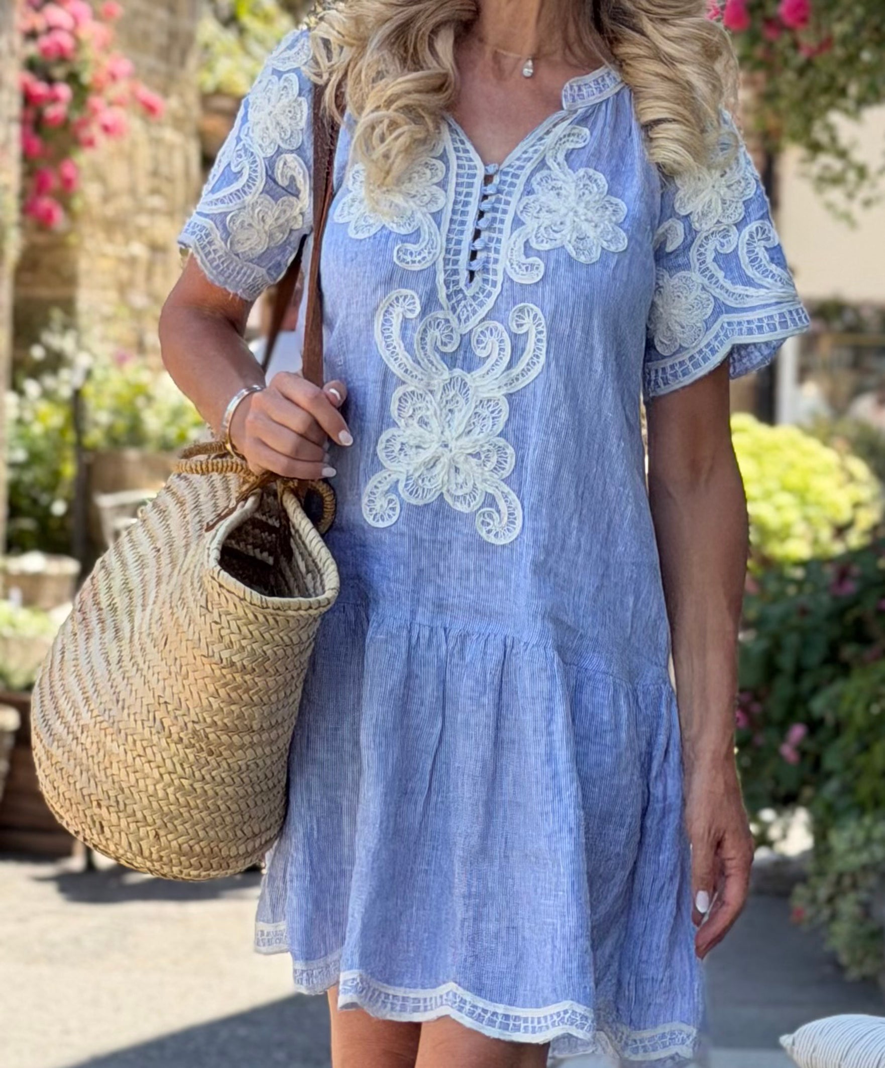 A woman wearing a striped blue short-sleeve Amaranta dress with white floral embroidery, holding a large woven straw tote bag in an outdoor garden setting.
