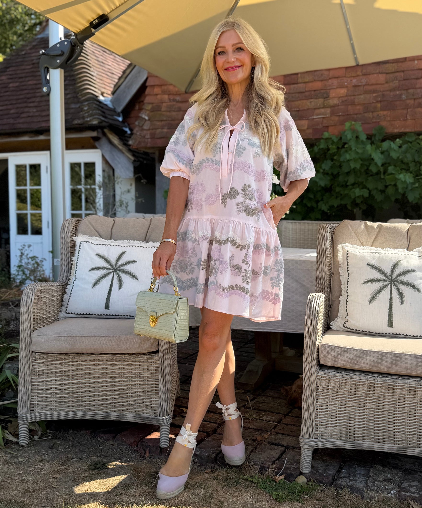 Influencer @seenonsarah stands outdoors in a garden patio setting wearing a light pink floral emb Oleander mini dress, holding a small handbag and wearing pale pink high espadrilles.