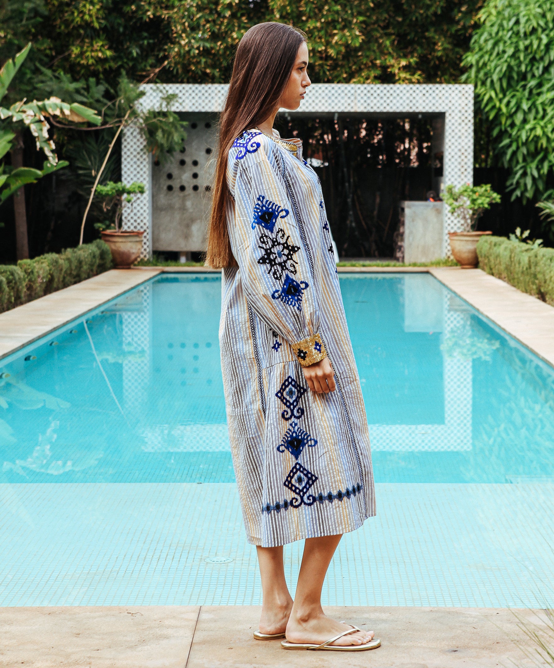 A side view of a model stood in front of a swimming pool wearing a Rose and Rose Temple striped dress with embroidery and sequin decoration.