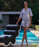 Woman in a floral embroidered shirt and shorts standing next to a modern black and white chair by a pool.